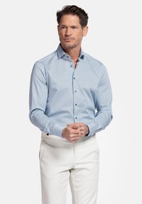 Chemise bleu clair à manches longues, à motif géométrique discret, avec un col pointu, associée à un pantalon blanc. Texture lisse.
