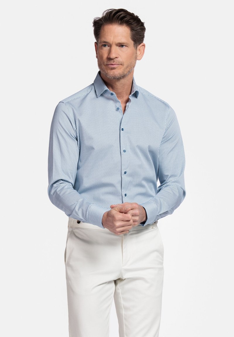 Chemise bleu clair à manches longues, à motif géométrique discret, avec un col pointu, associée à un pantalon blanc. Texture lisse.