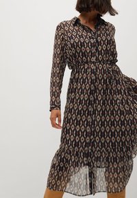 Vestido de manga larga y botonadura en tela oscura, con un patrón geométrico en crema y rojo. Incluye un dobladillo con volantes y una cintura ceñida con cinturón.