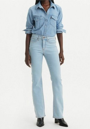 Femme portant une chemise en denim bleu clair avec manches retroussées, un jean bleu clair taille haute et des bottines noires à talons, debout sur fond blanc.