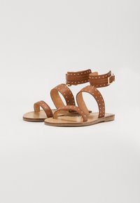 Brune lærsandaler med flere remme, naglede detaljer og en spænde lukning. Flad sål, åbent tådesign med en enkel, klassisk form.