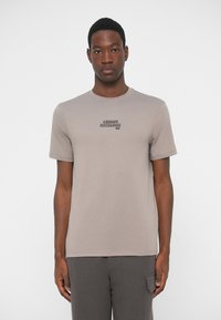 Young man wearing a taupe Armani Exchange short-sleeve T-shirt and dark gray pants, standing against a plain white background.