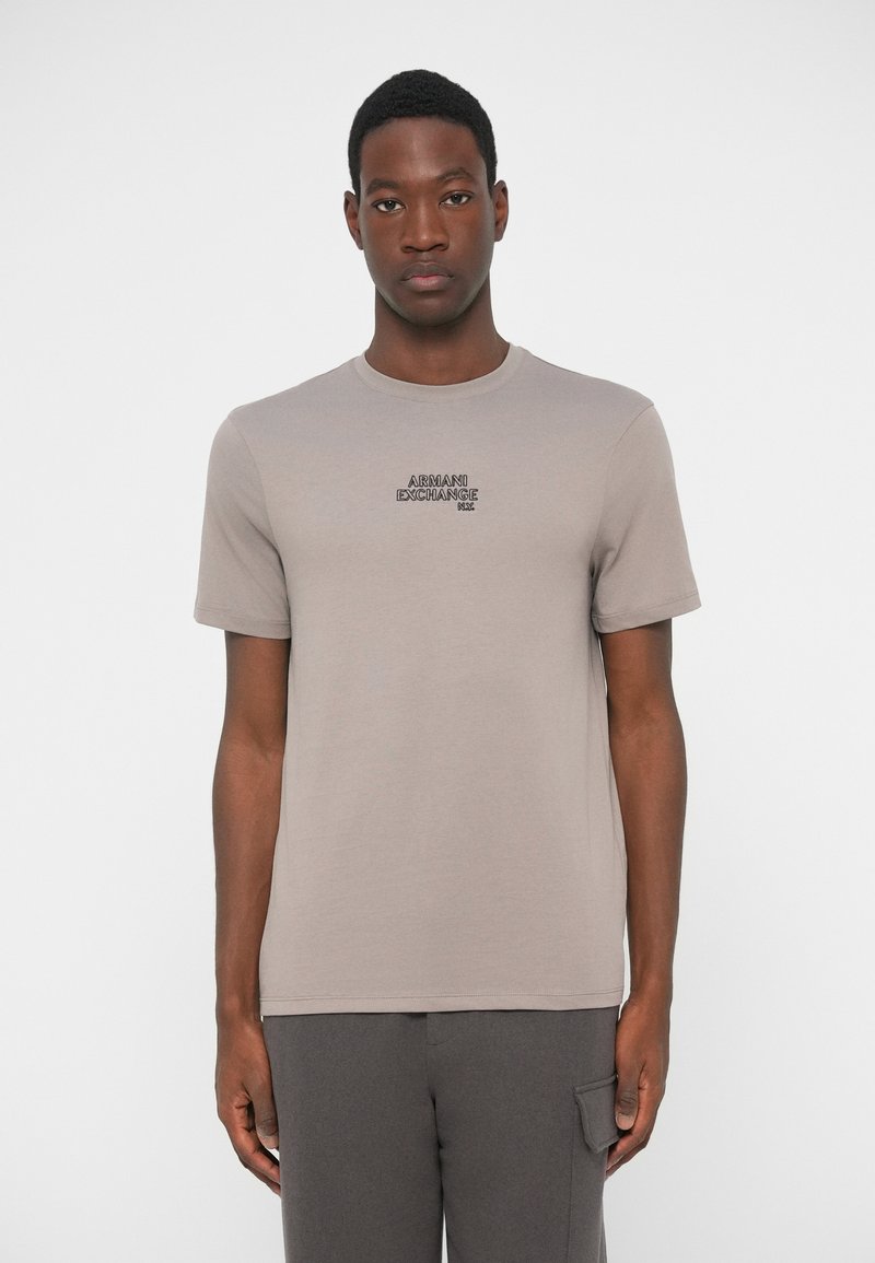 Young man wearing a taupe Armani Exchange short-sleeve T-shirt and dark gray pants, standing against a plain white background.