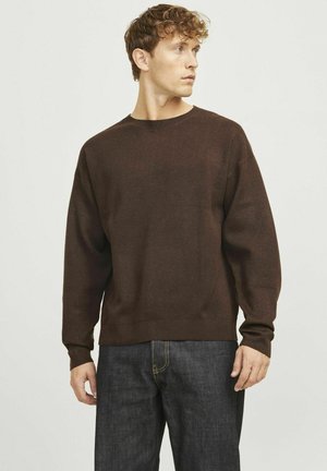 Suéter marrón con cuello redondo, hombros caídos y mangas largas. Textura suave y ajuste relajado, combinado con jeans de denim oscuros.