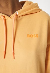 Primo piano di una persona che indossa un hoodie arancione chiaro con lacci abbinati e la parola "BOSS" stampata sul petto.