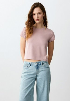 Woman with long wavy hair wearing a short-sleeve light mauve crop top and high-waisted light blue jeans, standing against a plain white background.