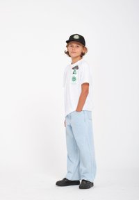 T-shirt blanc à manches courtes avec des motifs graphiques verts et violets ; jean baggy bleu clair ; casquette noire et chaussures noires complètent la tenue.