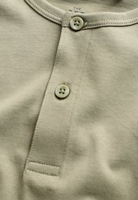 Khaki Baumwollhemd mit rundem Ausschnitt, zwei Knöpfen und einem kleinen gestickten Taschen-detail. Glatte Textur und weicher Stoff.