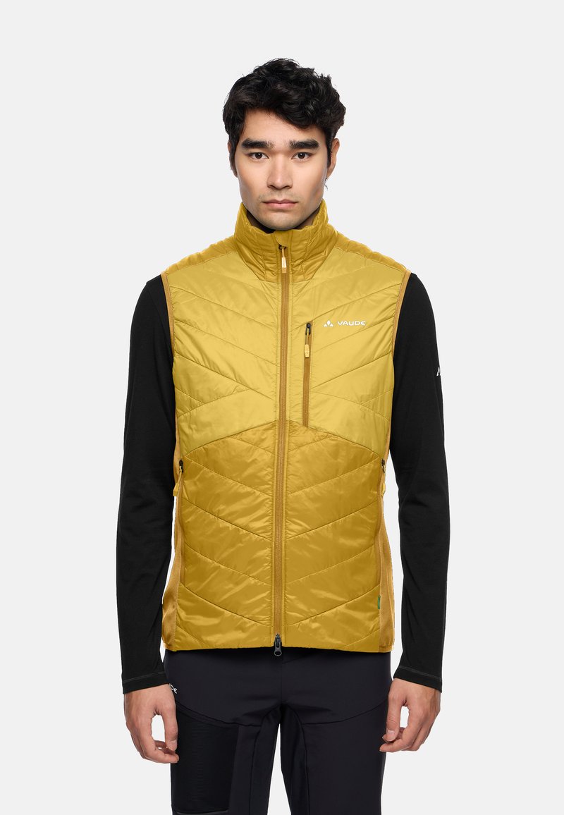 Gelbe gepolsterte Weste mit hohem Kragen, diagonaler Steppung und Seitentaschen. Getragen über einem schwarzen Langarmshirt.