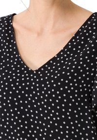 Blusa nera a V con un motivo a pois bianchi. Maniche corte, tessuto leggero con un aspetto testurizzato.