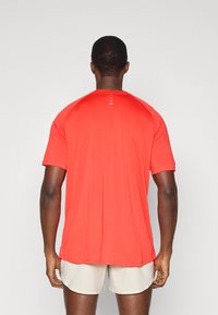 Κόκκινη αθλητική μπλούζα κοντό μανίκι με λεία υφή, που διαθέτει επίπεδη πίσω ραφή με δύο οριζόντιες ρίγες στον λαιμό. Φοριέται με ανοιχτόχρωμα σορτς.