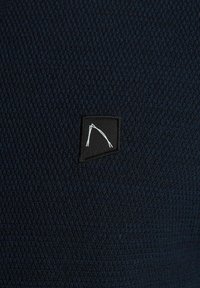 Tmavě modrá texturovaná látka s malou černou nášivkou s bílým geometrickým vzorem. Látka působí tkaně a má jemný vzor.