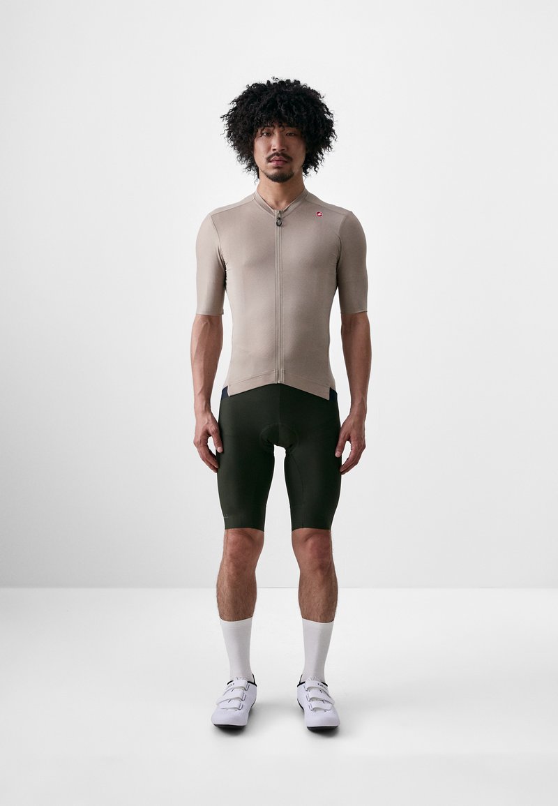 Jersey da ciclismo a maniche corte in beige con zip, abbinato a pantaloni corti da bici verde scuro. Modello che indossa scarpe e calze da ciclismo bianche.