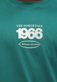 Tissu bleu sarcelle avec le texte blanc « THE NORTH FACE 1966 » et « EST. BERKELEY CALIFORNIA » imprimé sur le devant, stylisé avec des rayures horizontales.