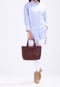 Burgundská kožená taška přes rameno s lesklým kovovým přívěskem. Má pevné rukojeti a hladkou strukturu, doplněnou modře pruhovanou košilí a bílými kalhotami.