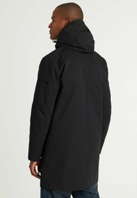 Juodas ilgas paltas su kapišonu, turintis lygų tekstūrą, matomus siūlių įpjovimus ir funkcionalias šonines kišenes. Sukurtas moderniam siluetui.