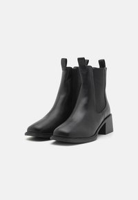 Botas de tornozelo em couro preto com painéis laterais elásticos, bico arredondado e salto bloco. Inclui puxadores para fácil calce. Textura suave.
