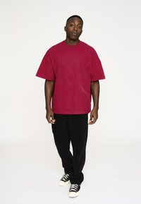 Rød oversized t-shirt lavet af bomuld, parret med sorte bukser. Personen bærer sorte og hvide sneakers. Enkel design uden mønstre.