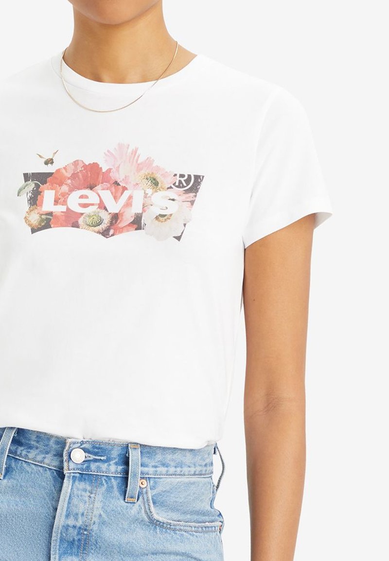 T-shirt bianco in cotone con grafica floreale e testo "Levi's" in lettere in grassetto. Maniche corte e vestibilità comoda. Pantaloni corti in denim visibili sotto.