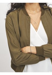 Femme portant des colliers en or superposés avec des croix, des croissants et des pendentifs, une veste bomber olive, un top blanc en V et un tatouage au poignet.