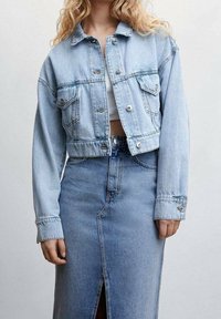 Giacca di jeans blu chiaro a maniche corte con due tasche sul petto e bottoni argentati, abbinata a una gonna di jeans a vita alta con uno spacco frontale.