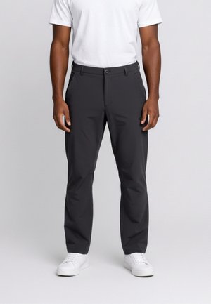 Uomo che indossa pantaloni neri a gamba dritta, scarpe da ginnastica bianche e una camicia bianca a maniche corte, in piedi davanti a uno sfondo grigio semplice.