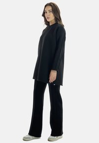 Felpa nera oversize con maniche lunghe, abbinata a pantaloni neri a gamba larga. Il materiale sembra morbido; dettaglio di cucitura notevole sulle maniche.