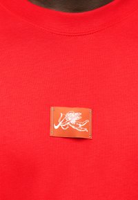 Κόκκινο πουκάμισο με ένα μικρό πορτοκαλί έμπλαστρο στο κέντρο του στήθους, που απεικονίζει ένα λευκό περίγραμμα ενός πετώντας χερουβείμ που κρατά ένα τόξο και βέλος.