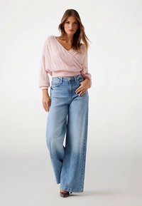 Pull tricoté rose pâle à manches larges, associé à un jean taille haute large bleu clair et des chaussures foncées. Textures lisses, style décontracté.