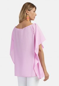Blusa rosa a vestibilità ampia con maniche larghe, orlo dritto e tessuto morbido; indossata con pantaloni bianchi, che mostrano un design fluido.