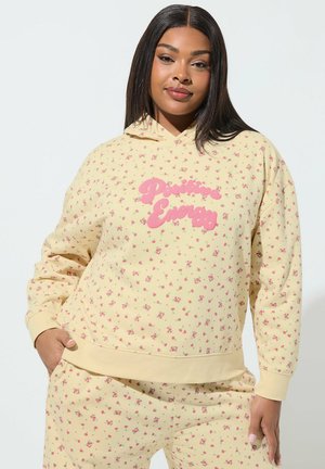 Frau trägt passenden hellgelben Hoodie und Hose mit kleinen rosa Blumen und dem Schriftzug "Positive Energy" auf der Vorderseite, posiert mit einer Hand in der Tasche.
