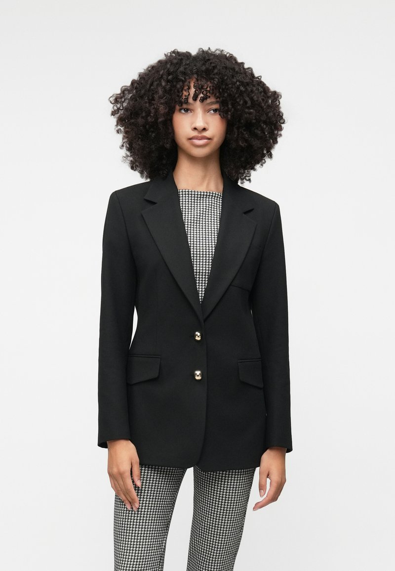 Mustat räätälöidyt bleiserit kultanappien kanssa, leikattu kaulus, kaksi etutaskua, päällä musta-valkoinen kuvioitu paita ja houndstooth-housut.