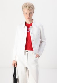 Jacheta din denim alb, cu două buzunare frontale, închidere cu nasturi și guler clasic, stratificată peste un tricou grafic roșu și pantaloni în culori deschise.