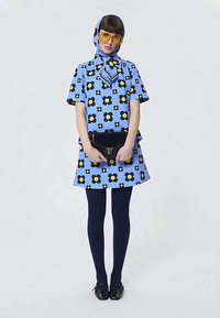 Kvinde i blå blomstret kjole og hovedscarf, gule solbriller, marineblå strømpebukser, sorte sko, holder en sort clutchtaske mod en hvid baggrund.