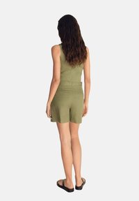 Rompers senza maniche verde oliva con pantaloni corti, presenta un corpetto aderente e una vita definita. Le tasche posteriori sono visibili; abbinato a sandali neri.
