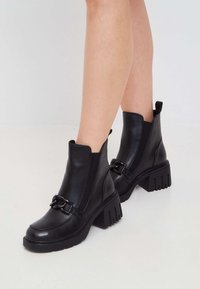 Botas de cuero negro hasta el tobillo con tacones de bloque y una suela texturizada, que cuentan con un detalle decorativo de cadena y paneles laterales elásticos para facilitar su uso.