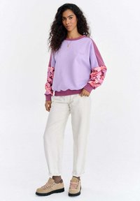 Lila dressipluus roosade lillemustriliste varrukatega ja ümara kaelusega, kombineeritud kreemikas värvi pükste ja pruunide matkasaabastega.