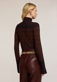 Maglione a collo alto lavorato a maglia con una base di un bordeaux scuro e un motivo a zig zag in toni più chiari, abbinato a pantaloni in pelle marrone e una cintura color sabbia.