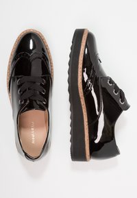 Chaussures en cuir verni noir à lacets avec une semelle en liège texturé, présentant des détails de brogue et des lacets noirs. Bout arrondi et semelle en caoutchouc épaisse.