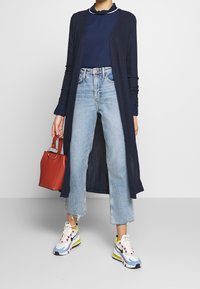 Długi granatowy kardigan w prążki założony na granatową bluzkę, jasnoniebieskie spodnie typu cropped oraz kolorowe sneakersy. Czerwona torebka w ręku dopełnia cały strój.