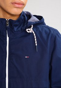 Marinblå jacka med dragkedja fram, dragsko huva och kontrasterande vit dragkedja. Har en broderad logotyp i rött, vitt och blått.