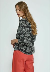Schwarze Bluse mit Blumenmuster und Puffärmeln, die weiße und rote Muster aufweist. Kombiniert mit korallefarbener weit geschnittener Hose für einen kontrastierenden Look.