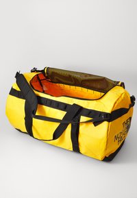 Bolsa de deporte amarilla hecha de material duradero, con correas negras, un bolsillo de malla en la parte superior y un interior naranja. Logotipo impreso en el lateral.