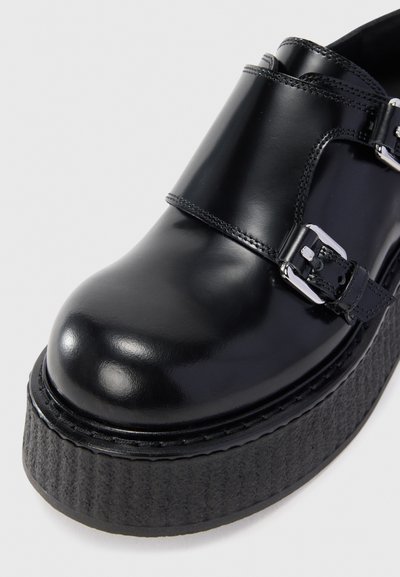 Black leather platform shoe with double silver buckles and thick textured sole on a white background.