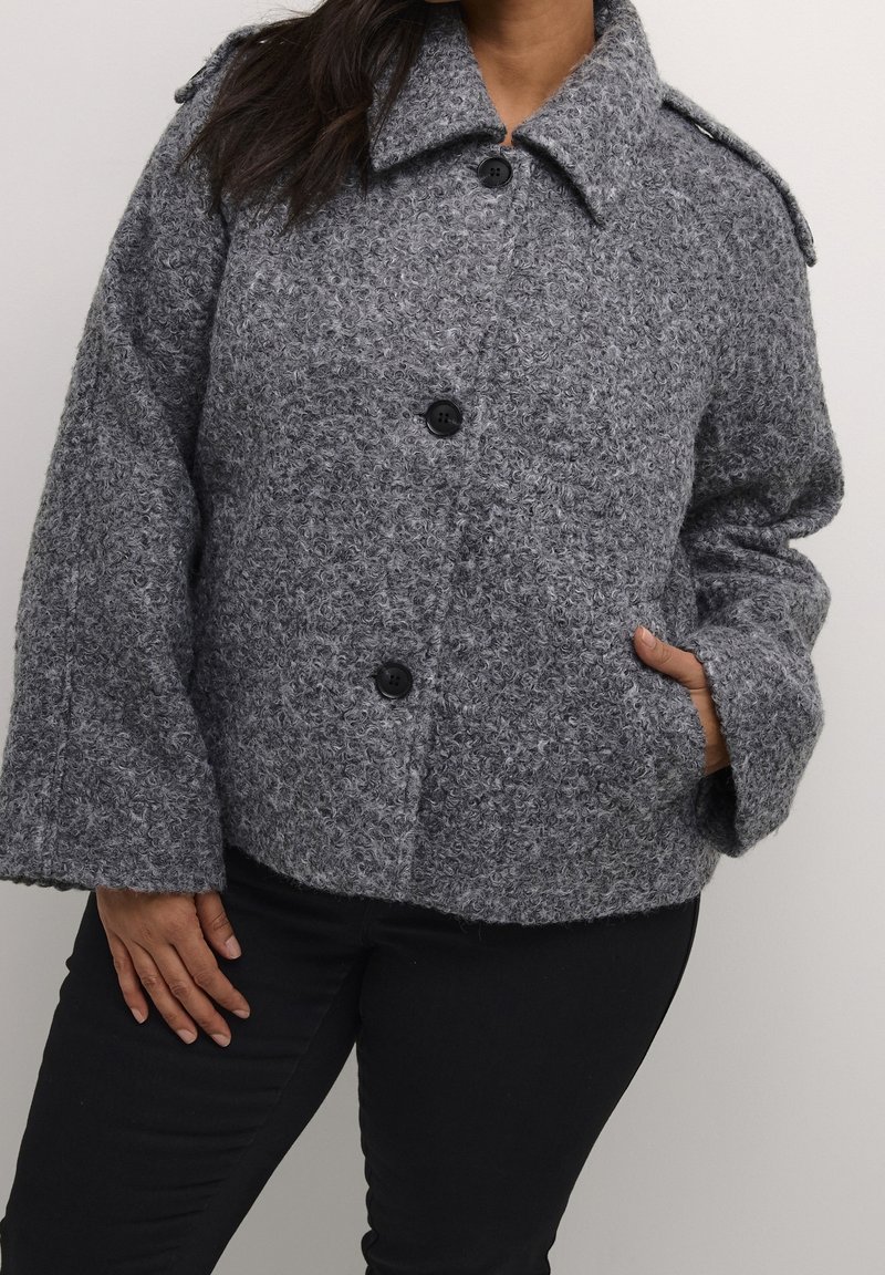 Grå tekstureret uldjakke med krave, fire sorte knapper og sidelommer. Har brede ærmer og en løs pasform.
