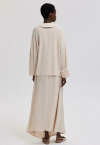 Beige, långärmad tröja med krage i ryggen, i kombination med en flödande, ankellång kjol, båda i mjukt, strukturerat tyg. Platta sandaler synliga.