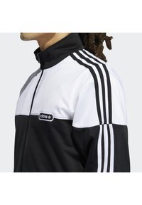 Svart och vit Adidas träningsjacka med dragkedja i kragen, med tre svarta ränder på varje axel och en liten logotyp på bröstet.