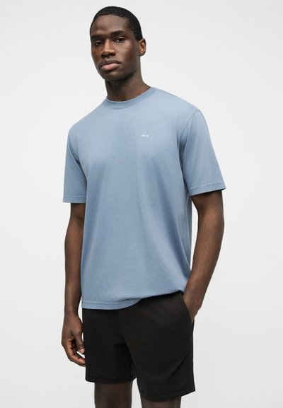 Homme portant un t-shirt à manches courtes bleu clair avec un petit texte blanc et un short noir, debout avec une main dans la poche.