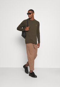 Maglione a maglia verde oliva con maniche lunghe, abbinato a pantaloni in velluto a coste marroni e scarpe nere stringate. Gli accessori includono occhiali da sole e una borsa nera.