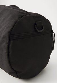 Sac cylindrique noir en tissu texturé. Il possède une fermeture éclair, un cordon de tirage et une poignée latérale pour un transport facile.